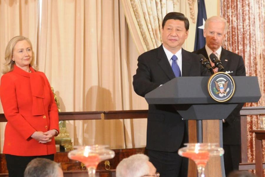 Chinese President Xi Jinping standing at a podium with the US Seal on the front, with Joe Biden behind him and Hilary Clinton to his left dressed in a red pants suit.