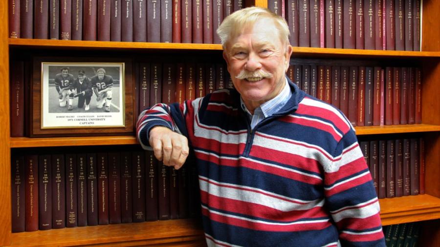 David Meehl, with a white mustache and striped shirt, stands smiling adn leaning on a bookcase.