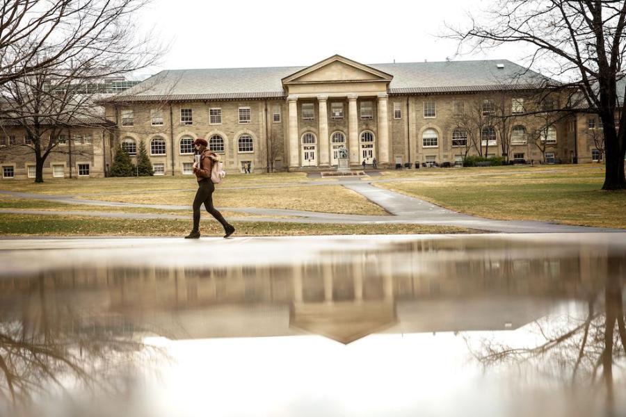 person walks past a puddle that is reflecting a campus building