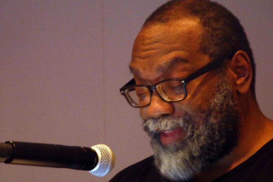 Fred Moten, with a mostly white beard and black glasses in front of a microphone