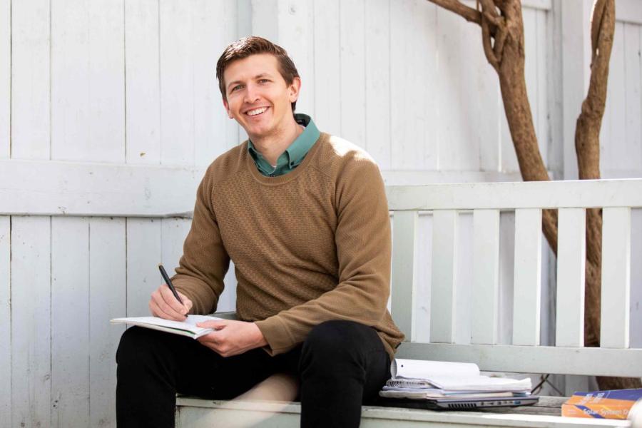 J.J. Zanazzi sitting on a white wooden bench underneath a leafless tree, writing in a notebook; smiling.