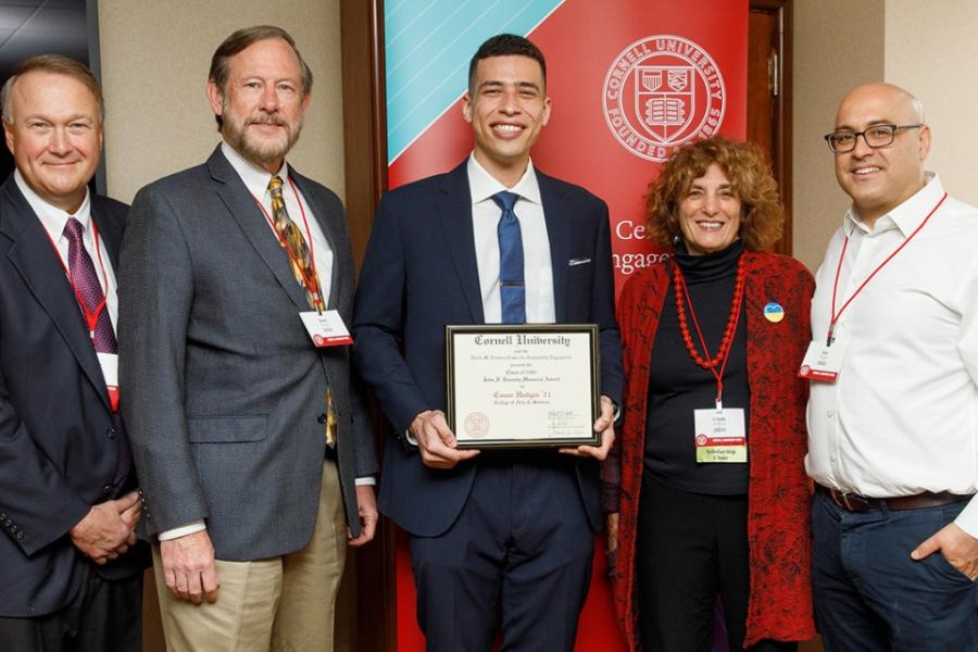 Conor Hodges in suit and tie, smiling and holding his award plaque, flanked by other alumni.