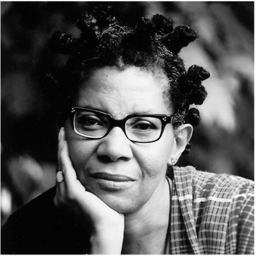 M. NourbSe Philip wearing glasses and a checkered top, leaning on her hand.