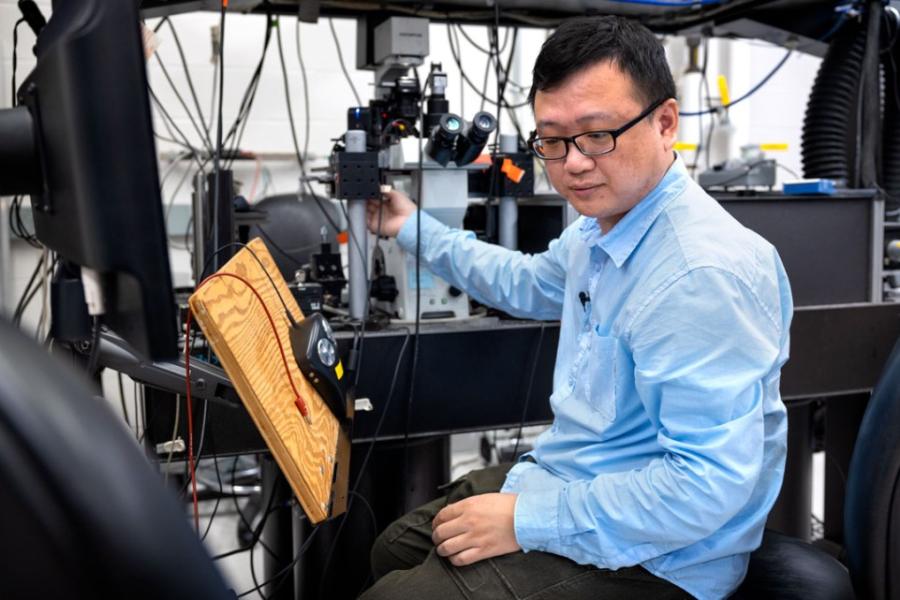 Wei Wang, in a blue shirt and black plastic-framed glasses, sits in a lab looking at an instrument while he adjusts another instrument with his right hand.