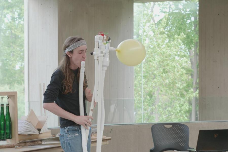 J Nation blowing on an instrument made out of long white pipes, with a yellow balloon attached