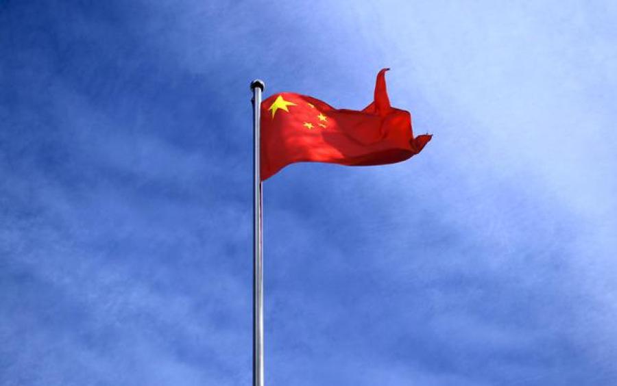 The Chinese flag, red with a circle of gold stars, waving on a flagpole against a blue sky. 