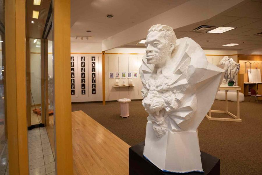 An African man's head with a ray-like collar above the face of a roaring lion with other artwork from the Sculpture Shoppe exhibition in the background.