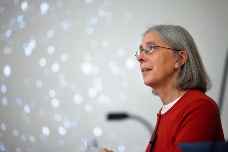 Martha Haynes with glasses, shoulder-length gray hair in a red top, with blurred stars on screen behind her
