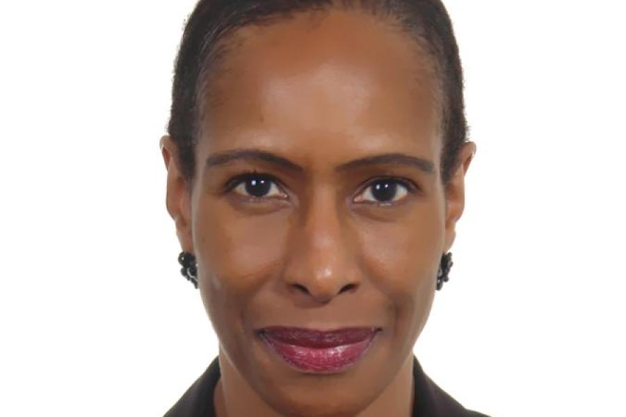 Ann Simmons smiling, with very short hair, red lipstick, earrings and a black jacket over a black top.