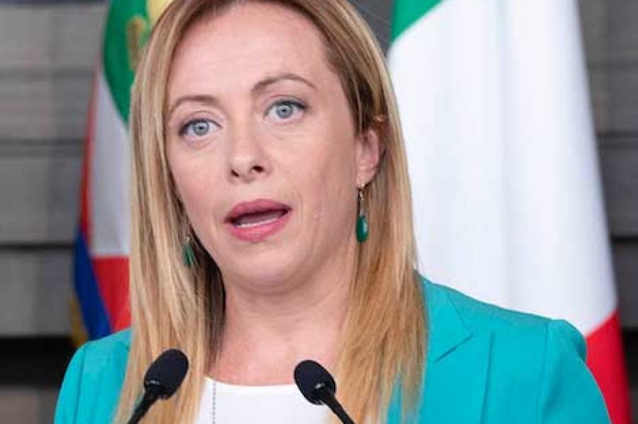 Person in blue jacket speaking in front of red, white and green flags