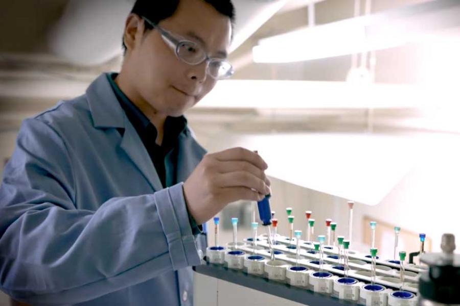 Person in lab coat and safety glasses, placing a tube in a rack