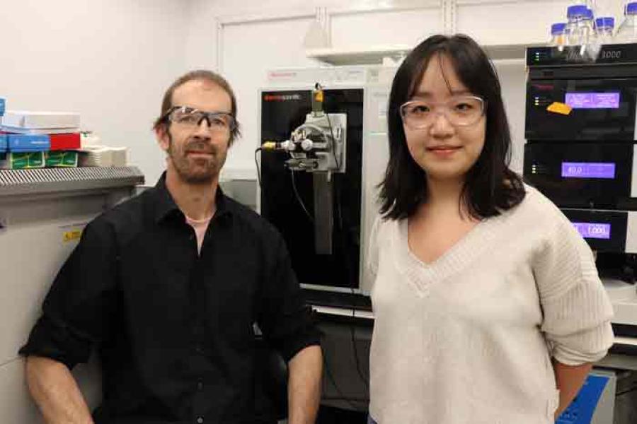 Frank Schroeder in glasses, beard and black shirt in front of lab equipment and next to woman with long black hair, white shirt and glasses.