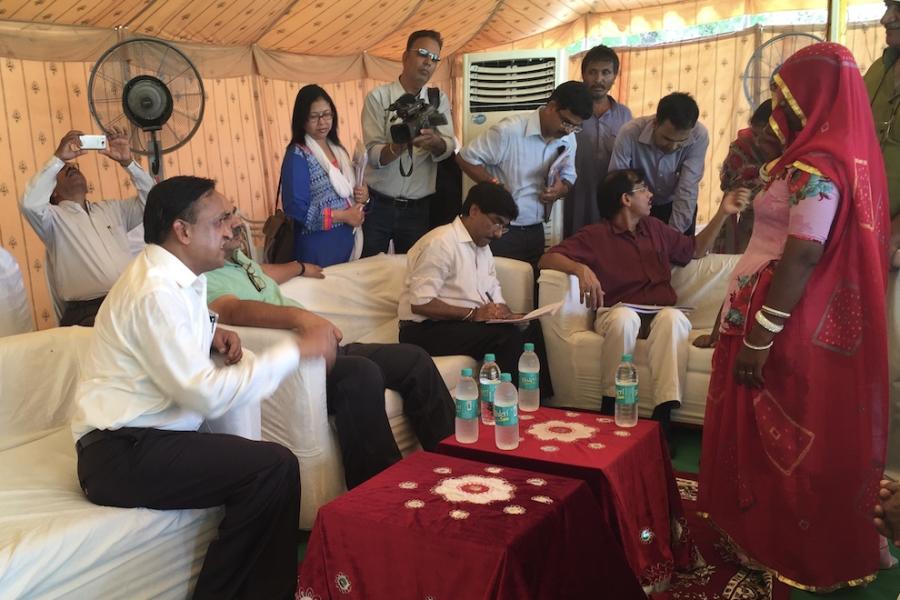 Among several people in a tent, a woman in red traditional clothing faces three officials in white shirts and black trousers, sitting on a couch