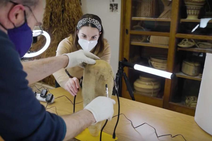 Two people wearing gloves work with football-sized museum object