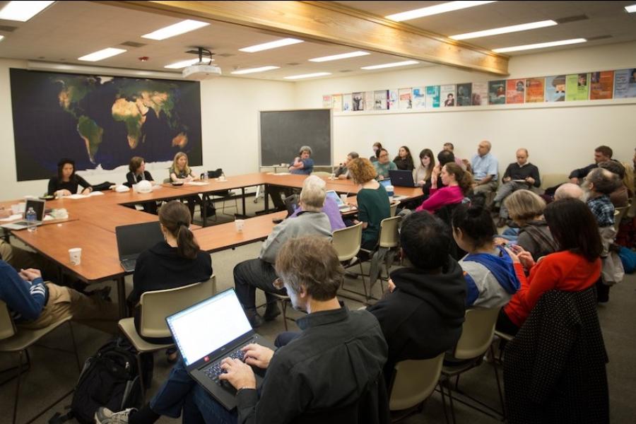 A few dozen people sit around a large square of tables in a room decorated with maps