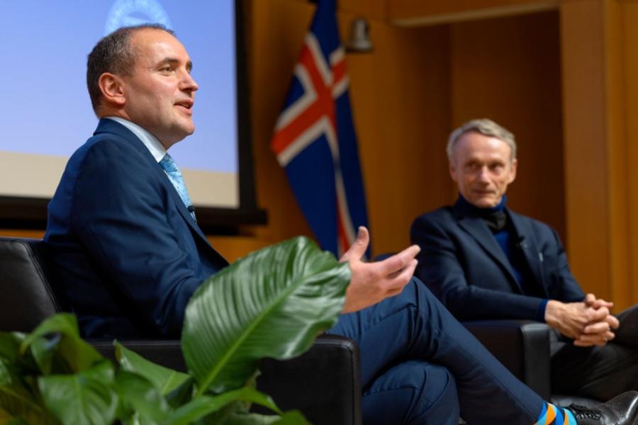 Two people wearing suits speak, seated on a stage among plants
