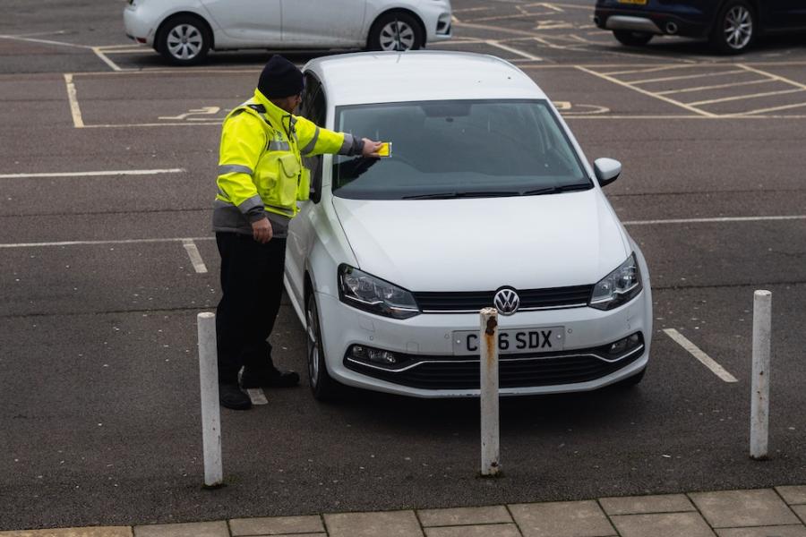 Person wearing a bright yellow jacket places a ticket on a car windshield