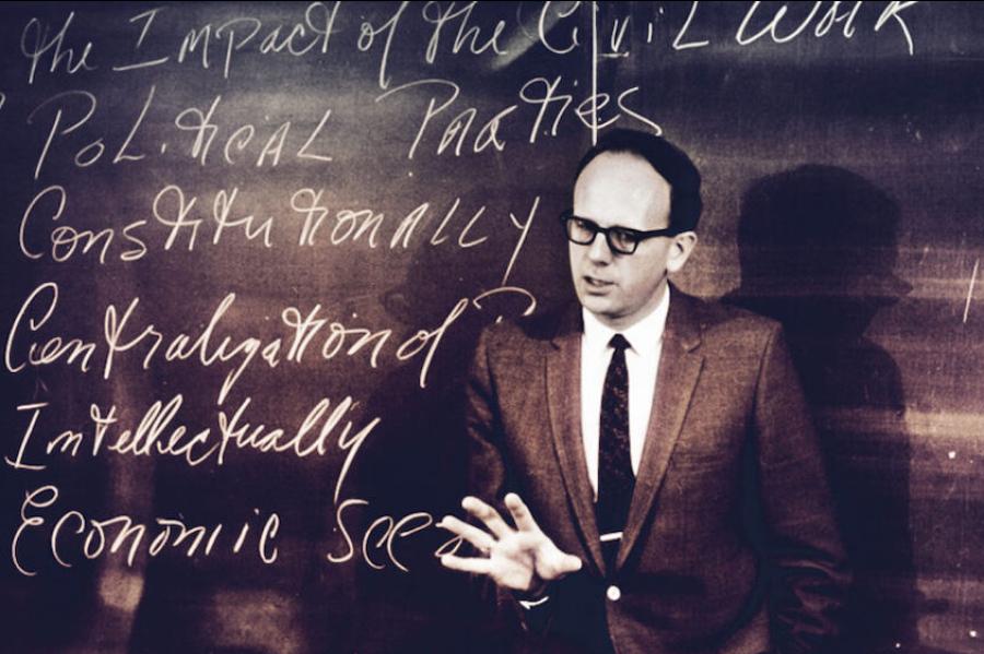 Black and white historical photo of a person in jacket and tie speaking authoritatively in front of a chalk board