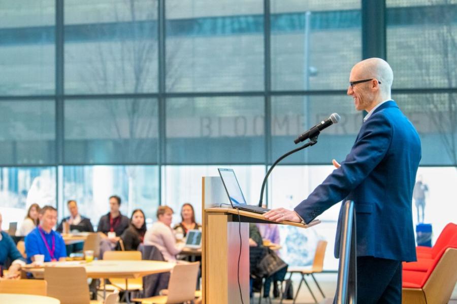 Person speaks to a group from a podium with a microphone: large windows in the background