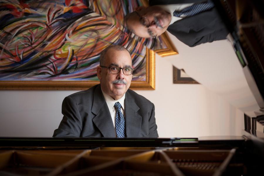 Person wearing a suit, sitting at a piano with a colorful painting in the background