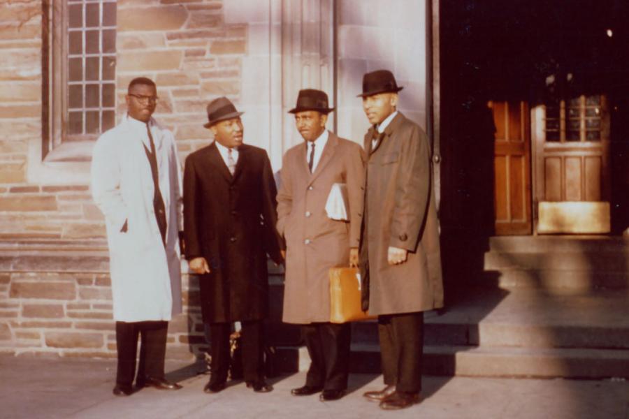 Four people stand in front of a building, wearing dress coats and hats