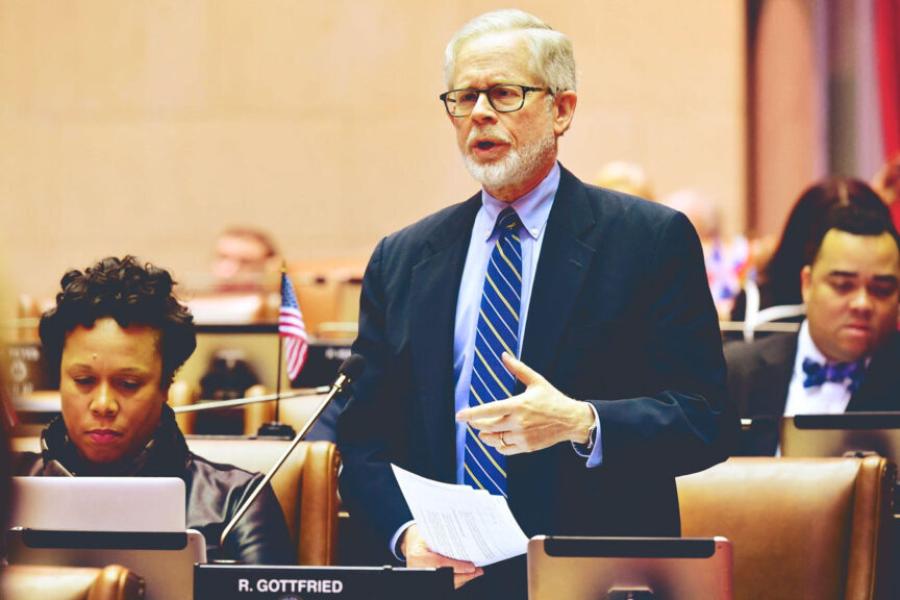 Person wearing a suit standing behind a desk, speaking seriously