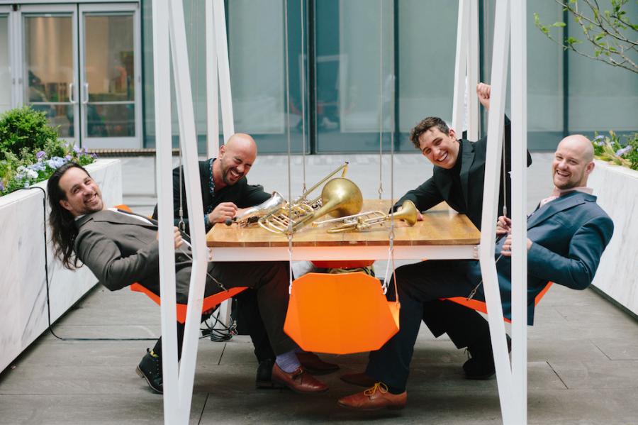 Four people sitting around a table that has musical instruments on it: a saxophone, a trombone and a trumpet