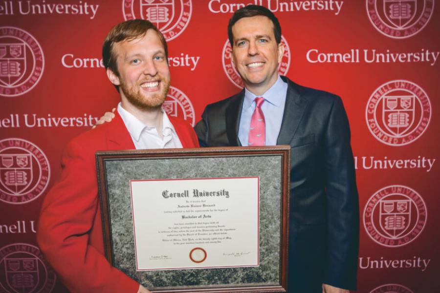 Two people stand in front of a red backgroun, holding a framed diploma