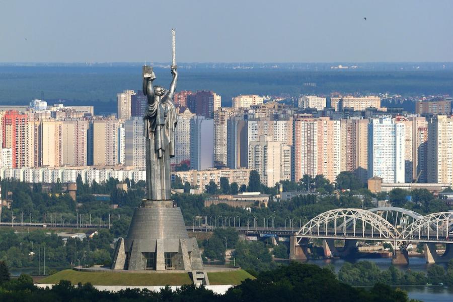 Tall monument in the shape of a figure holding a sword; city buildings in the background