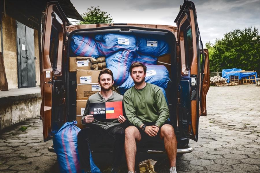 Two people sitting in the back of a van with doors open, showing boxes and bags