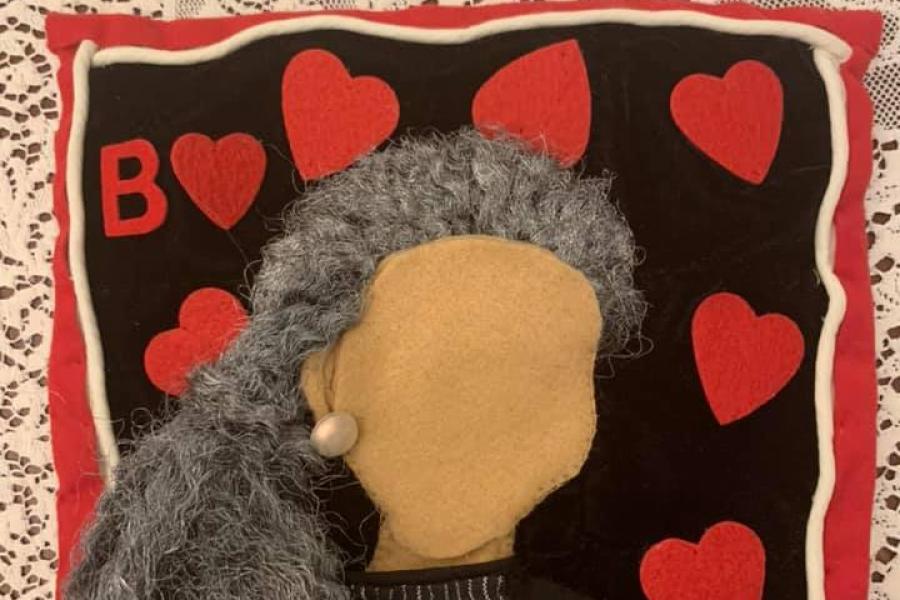 A red bordered rectangle made of felt with red hearts on it and a blank-faced brown-skinned woman with one pearl earring and long gray hair