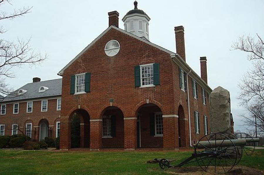 A red brick building with a white painted cupola on top with a weather van, with three large archways in front and a side building. A cannon sits in front.