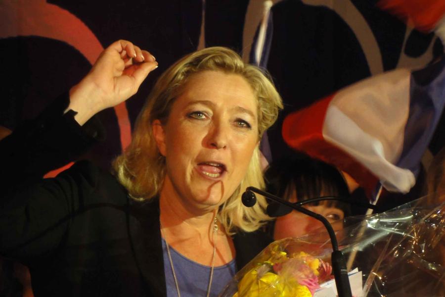 Marine Le Pen with sholuder-length blonde hair and jacket, with hand upraised in the midst of a speech, with French flag in bakcground