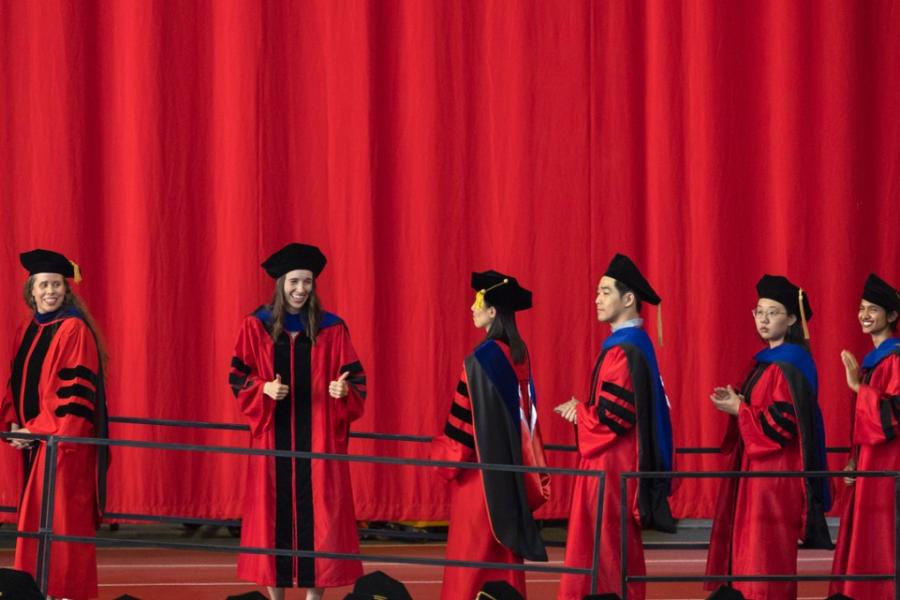 Several people in long red robes and black caps walk in a line against a red background; one turns to give a thumbs up