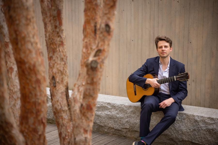 Person sitting on a stone wall, holding a guitar near trees