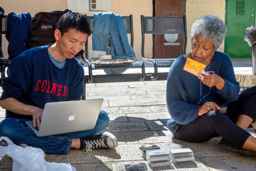 Two people sit on the ground; one types on a laptop, the other holds a compact disc