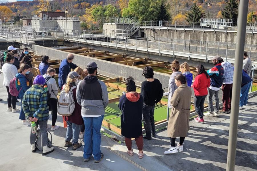 Students standing around an open wastewater treatment facility, which looks like a big cement rectangle with green water and wooden separaters.