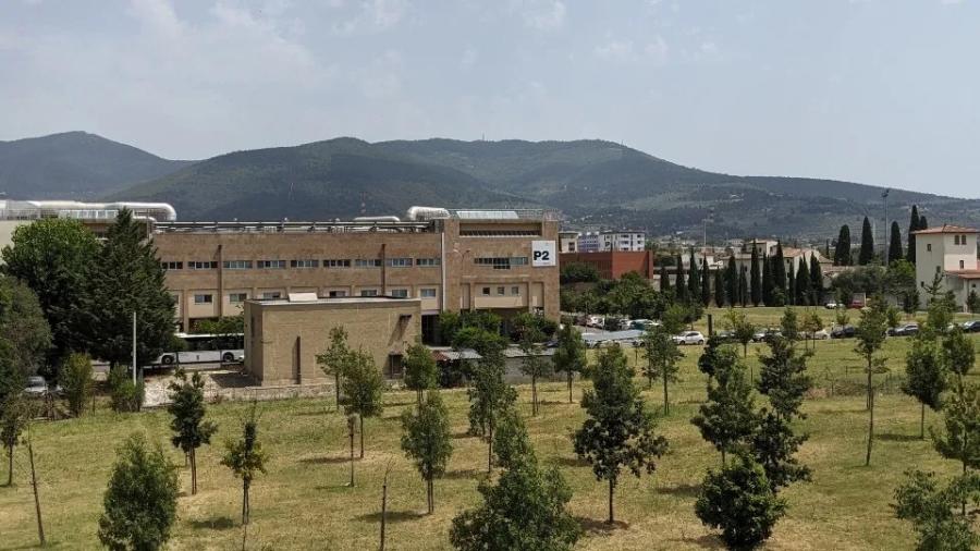 A long building with a row of windows and silver pipes coming out of the roof with scattered trees in a field in front. The Tuscan hills loom behind the building.