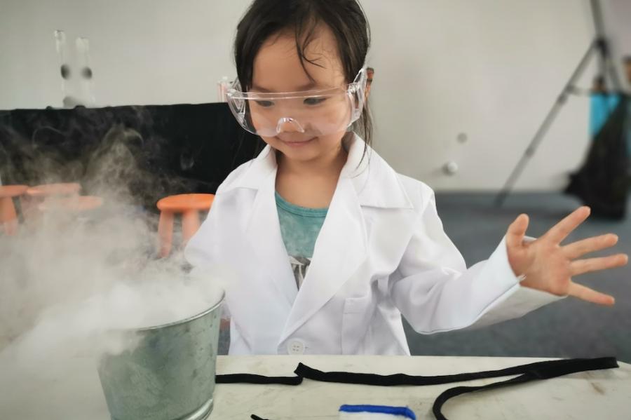Small person wearing safety goggles and a white lab coat, smiling as smoke pours out of a beaker on a lab bench