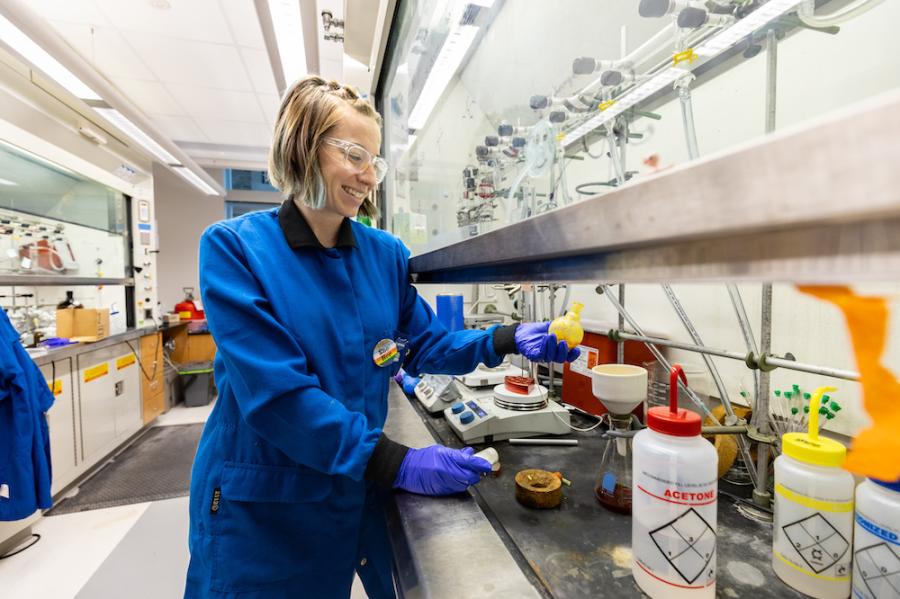 Person in blue lab coat, standing at a counter full of instruments and bottles