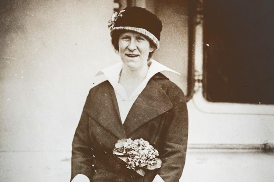 Black and white photo from 1914: a woman in a dark suit and hat highlighted by flowers stands on a wooden dock