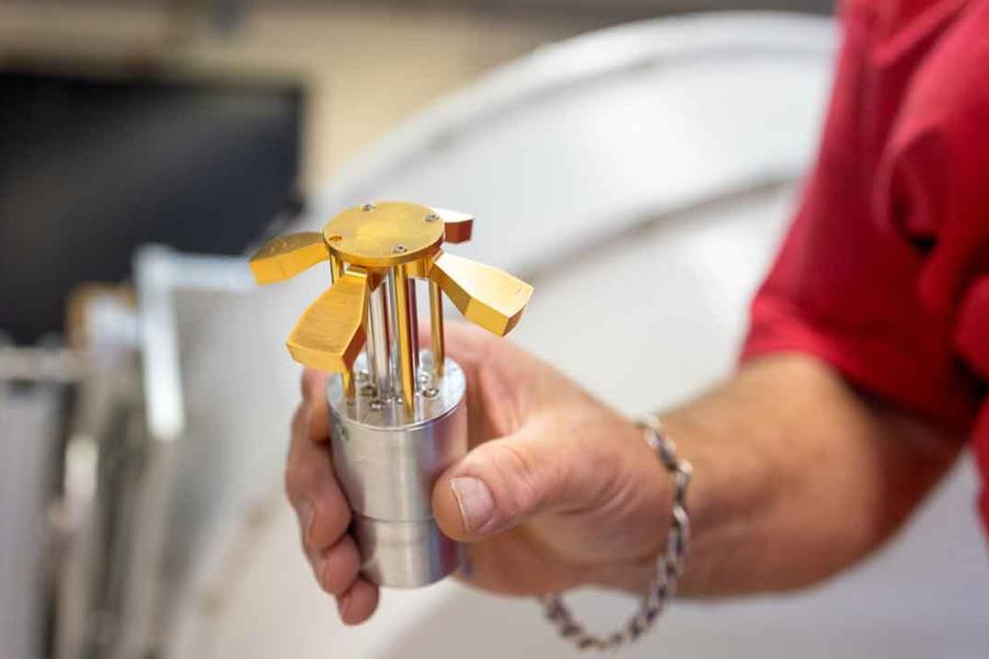 A hand holds a round silver-colored metal piece with a gold-colored whirligig on top, with four wings and a round center
