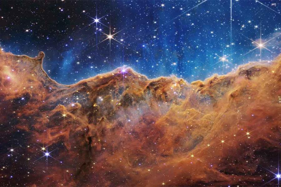 A field of stars in the background and in the foreground a colorful cliff-shaped mass of cosmic gases.
