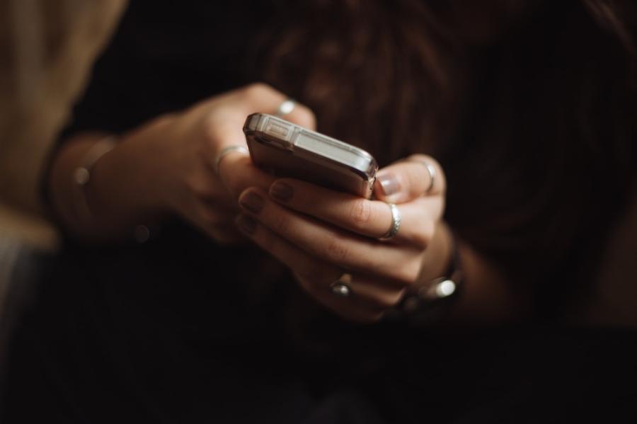 Two hands (manicured, wearing silver rings) hold a smart phone against a dark backgroun