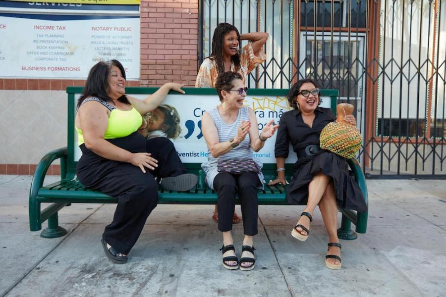 Three people sitting on a city bench with one standing behind; they are laughing together