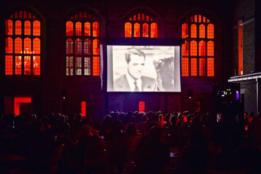 Movie screen outdoors, showing a black and white still of Jimmy Stewart, with red-lit windows behind it. 