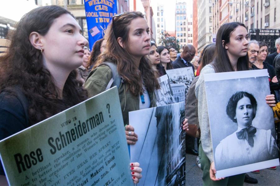 People in a crowd look thoughtful and carry signs depicting women from around 1911