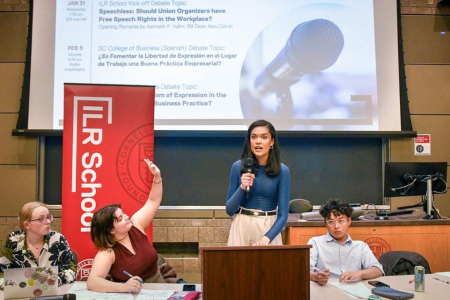 Person stands with microphone, speaking intently while three others listen