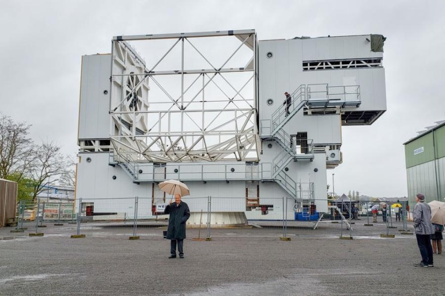Fred Young in raincoat, holding umbrella, in front of huge metal structure of telescope, with a handful of other people with umbrellas watching him.