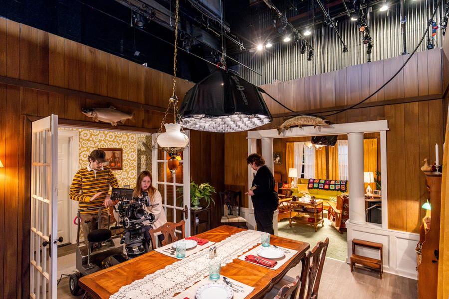 three people working in a film set that looks like a mid-century living room. The fly space of a theater is visible above the room's walls